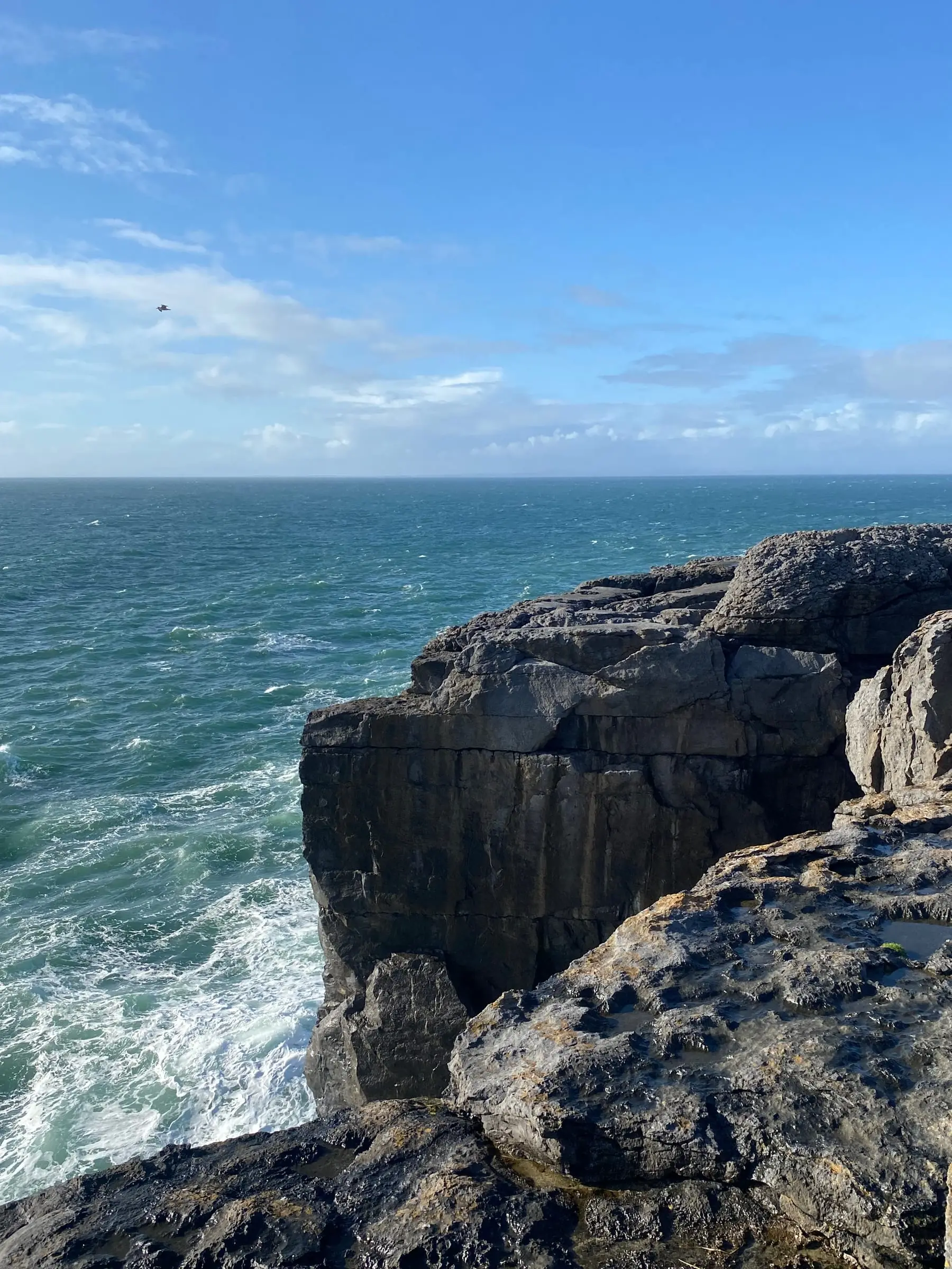 Layered grey rock ledges at the edge of the sea with green-blue water breaking below under a blue sky