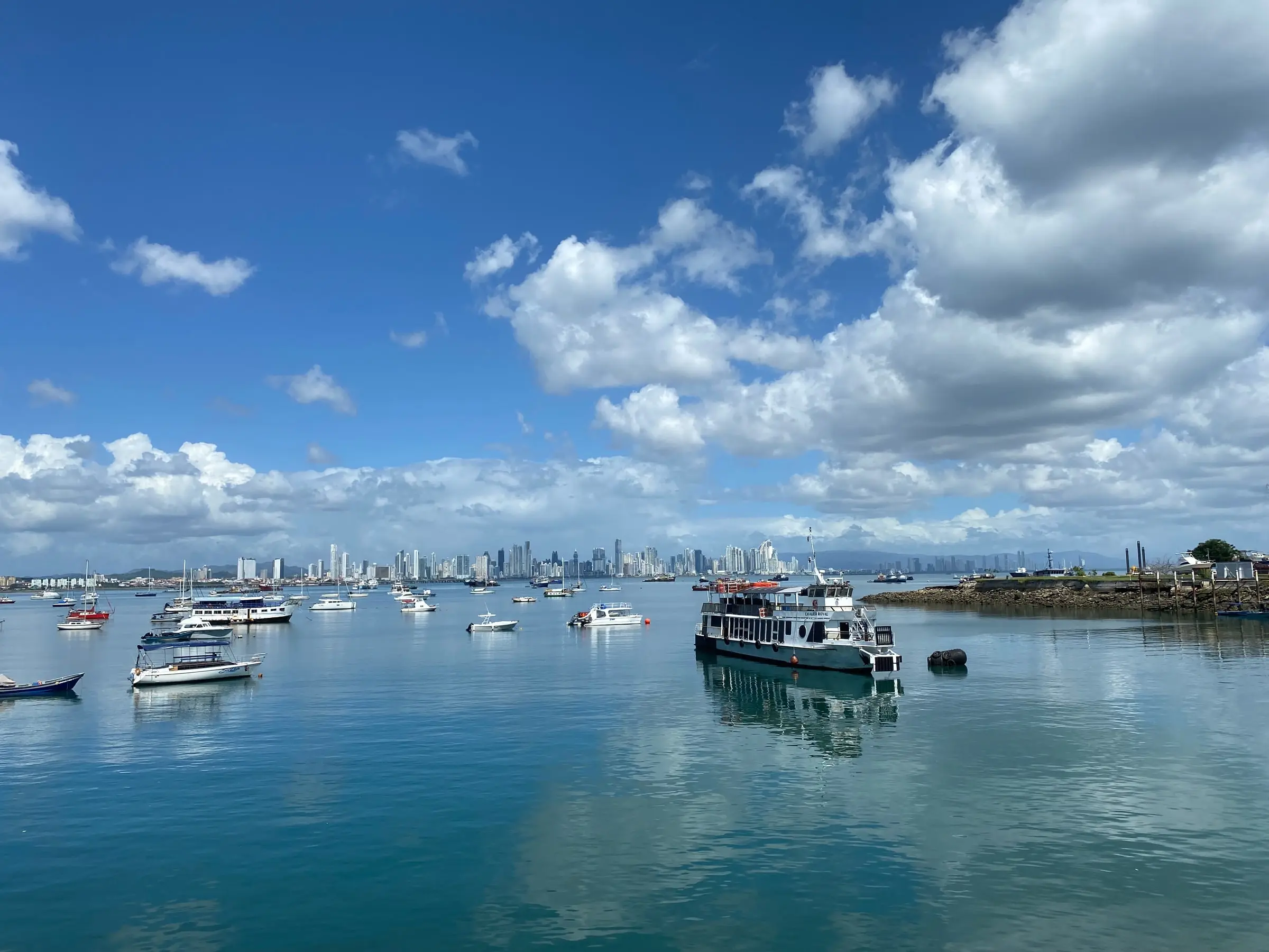 Boats at anchor on calm turquoise water under big clouds, the Panama City skyline in the distance