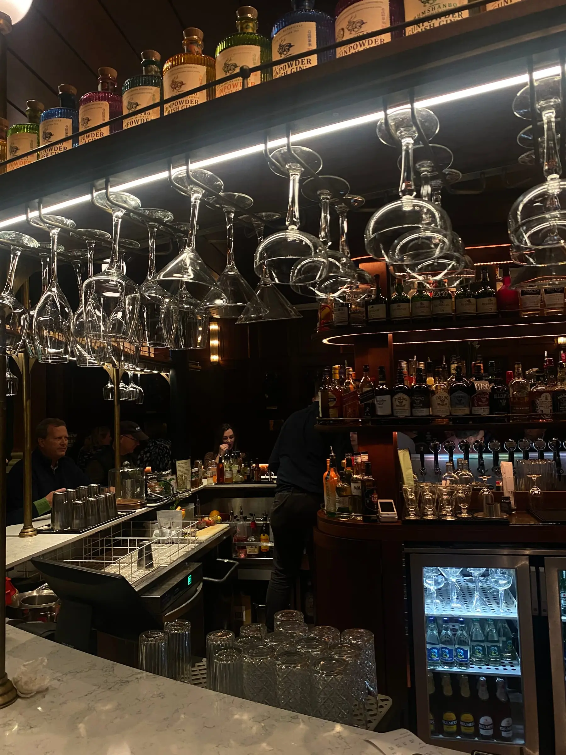 Rows of wine glasses hanging upside down above a bar, bottles and a lit fridge behind the counter