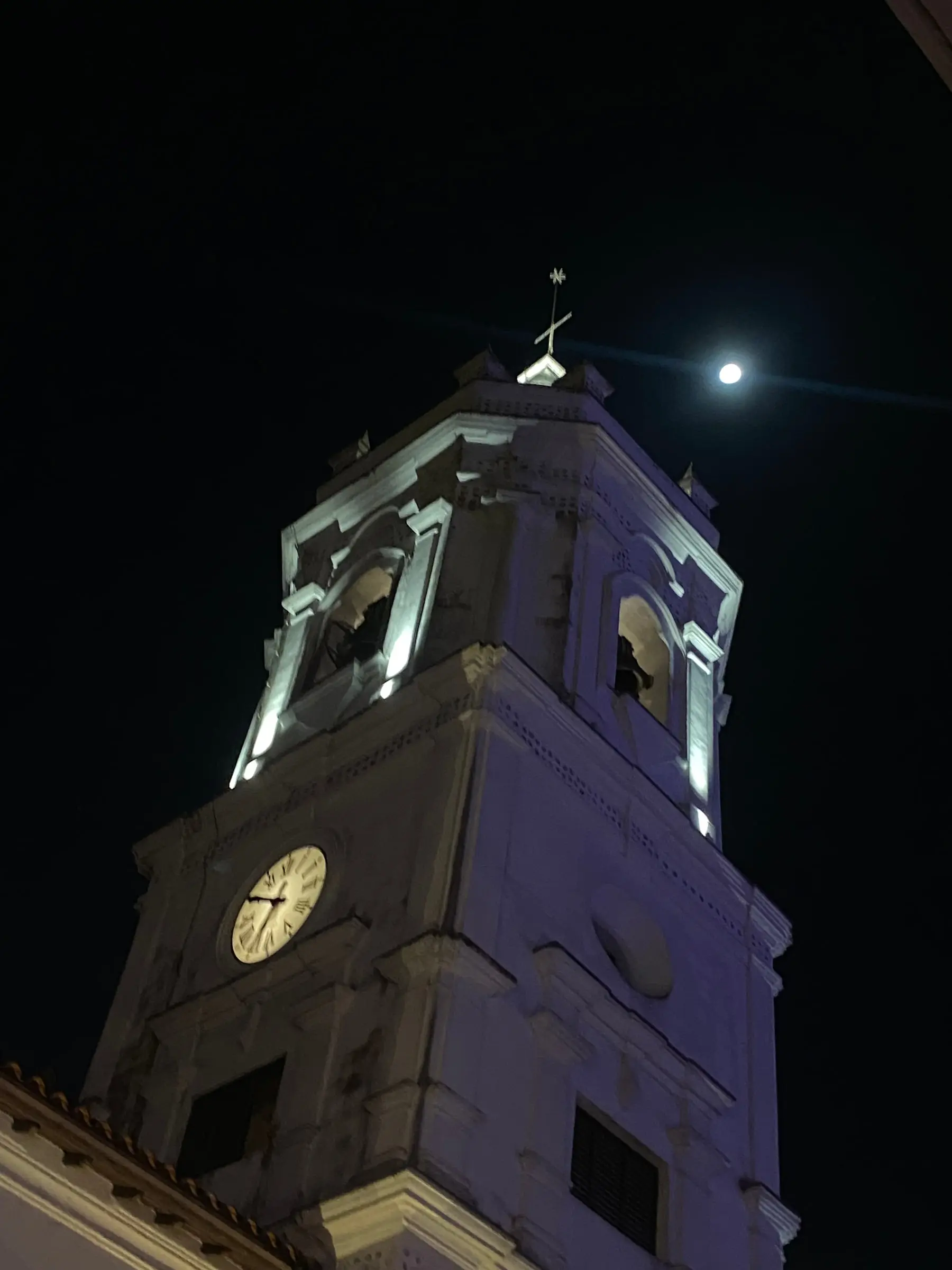 A floodlit white bell tower with a clock against the night sky, a bright moon beside its cross