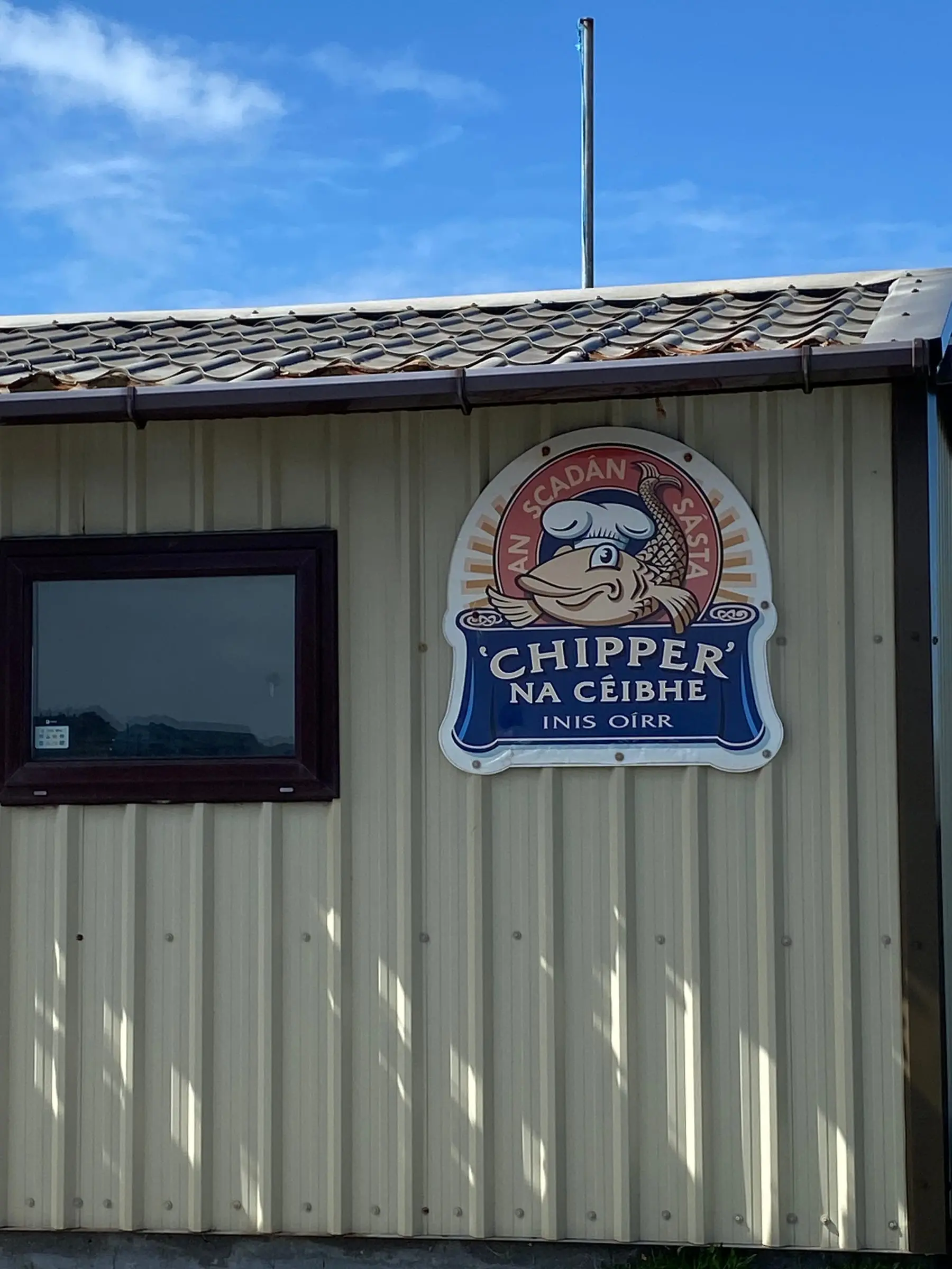 A corrugated hut with a round sign showing a cartoon fish chef and the words Chipper na Céibhe, Inis Oírr
