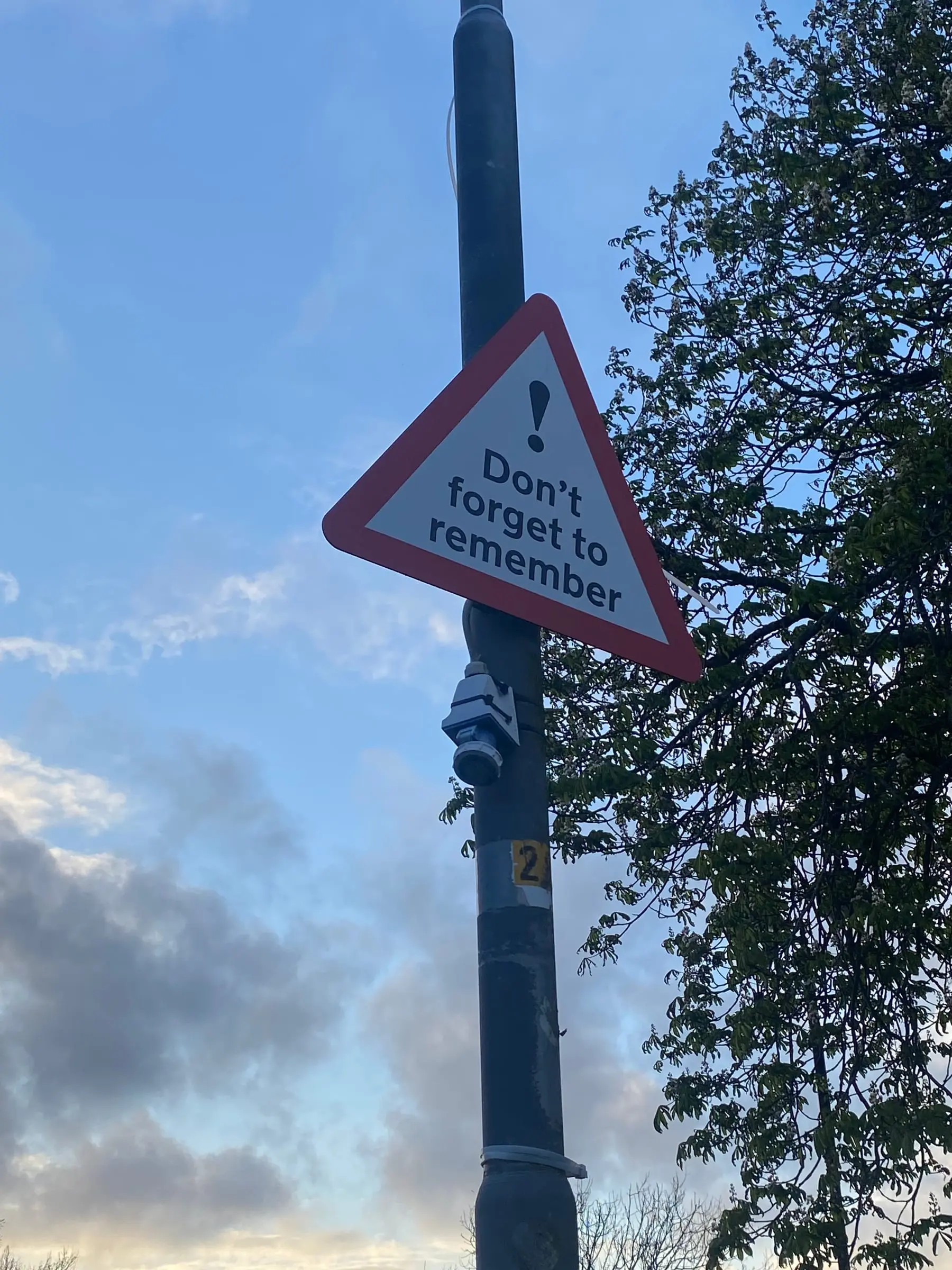 A red-bordered triangular warning sign on a pole reading Don't forget to remember, against a dusk sky and a leafy tree