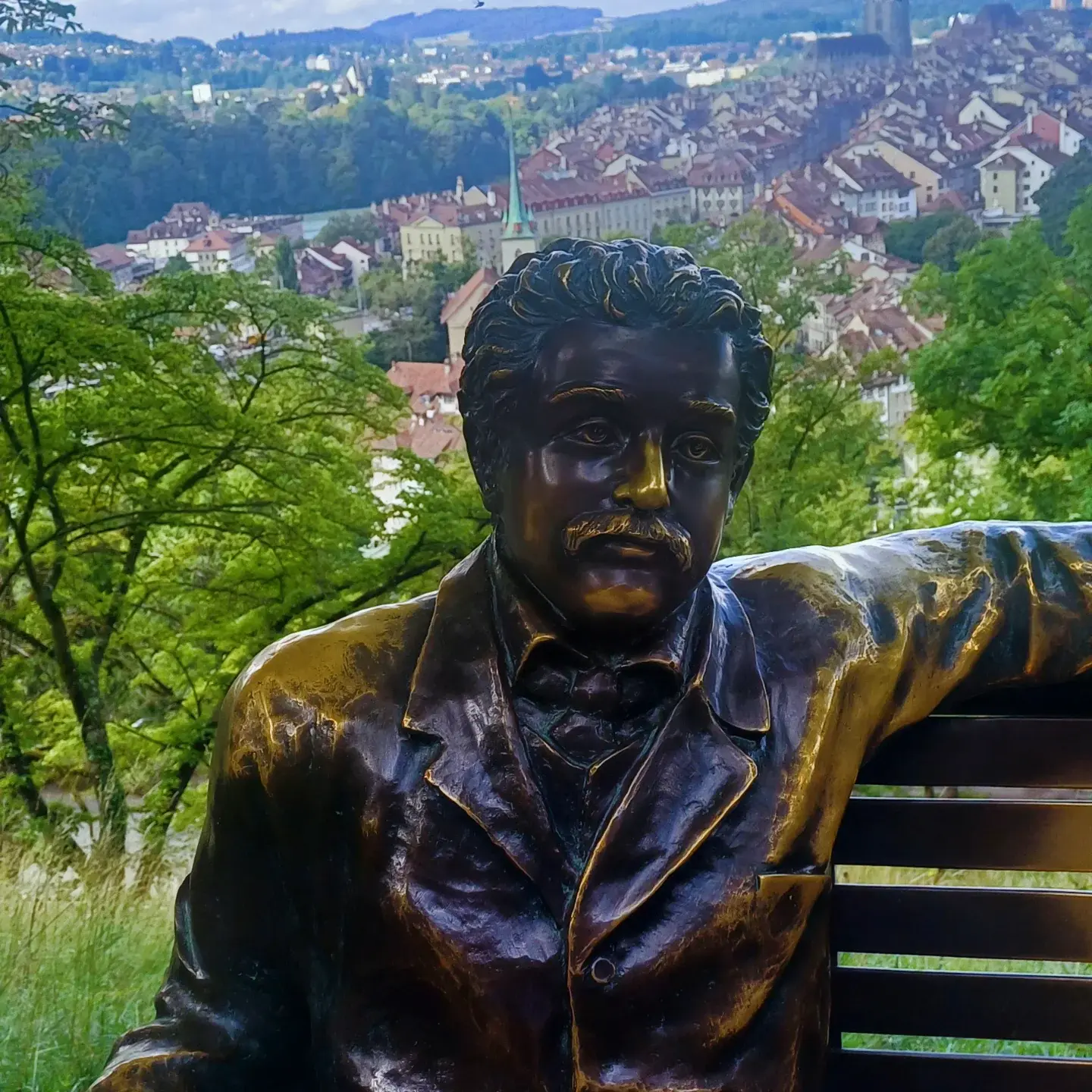 A bronze statue of Albert Einstein seated on a bench, the city of Bern visible through the trees behind