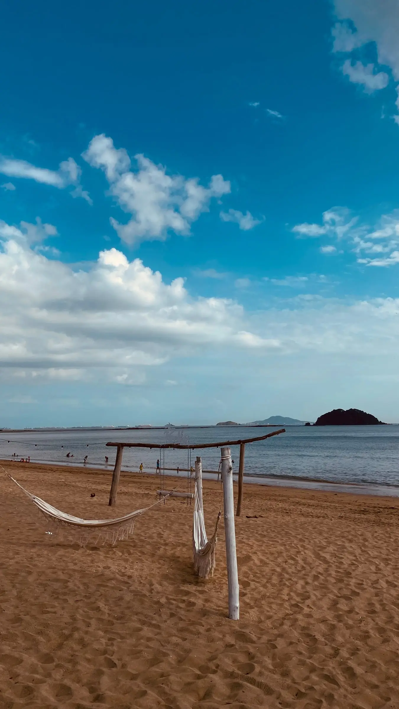 A white hammock strung between wooden posts on an orange sand beach, clouds over the sea and a dark islet