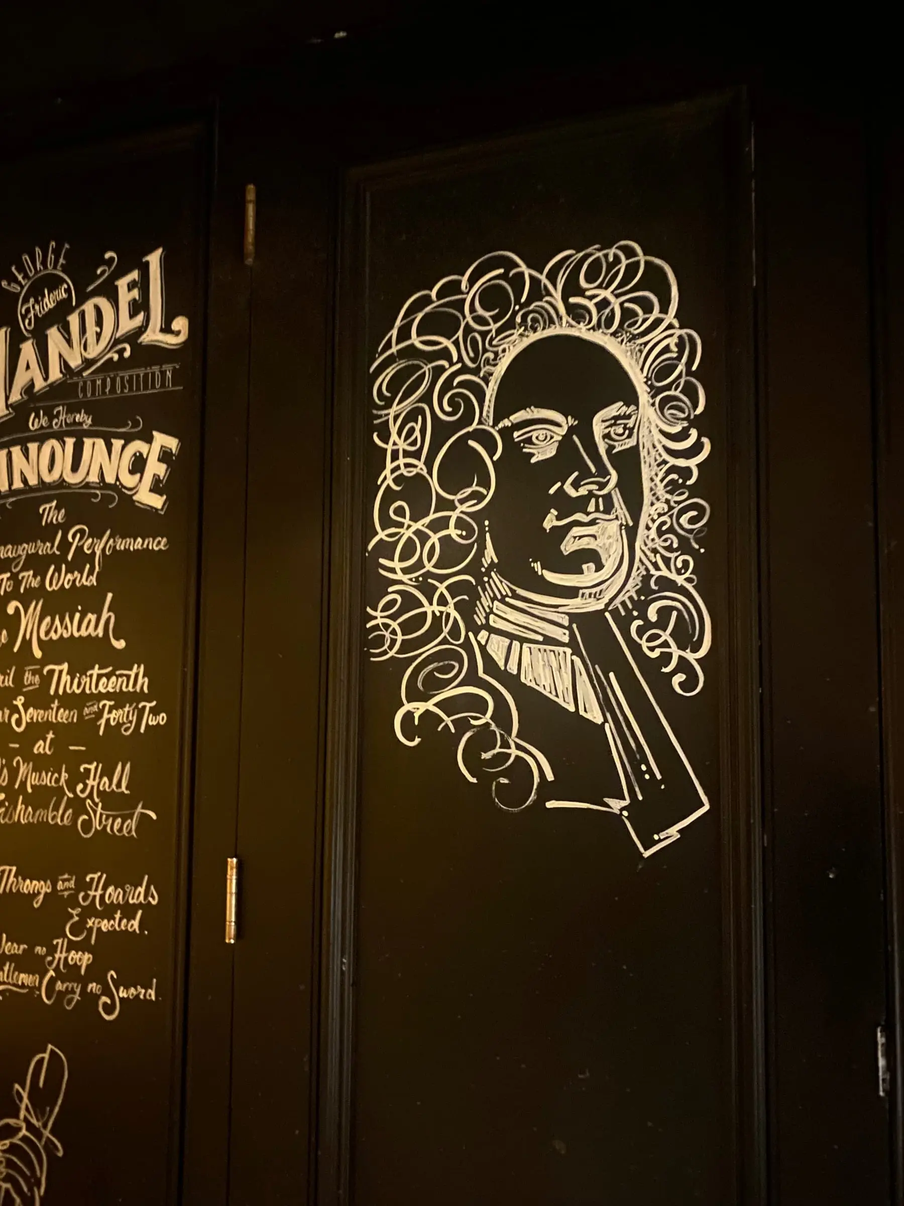 A chalk drawing of a man in a long curled wig on a black wall, next to a chalkboard announcing Handel's Messiah