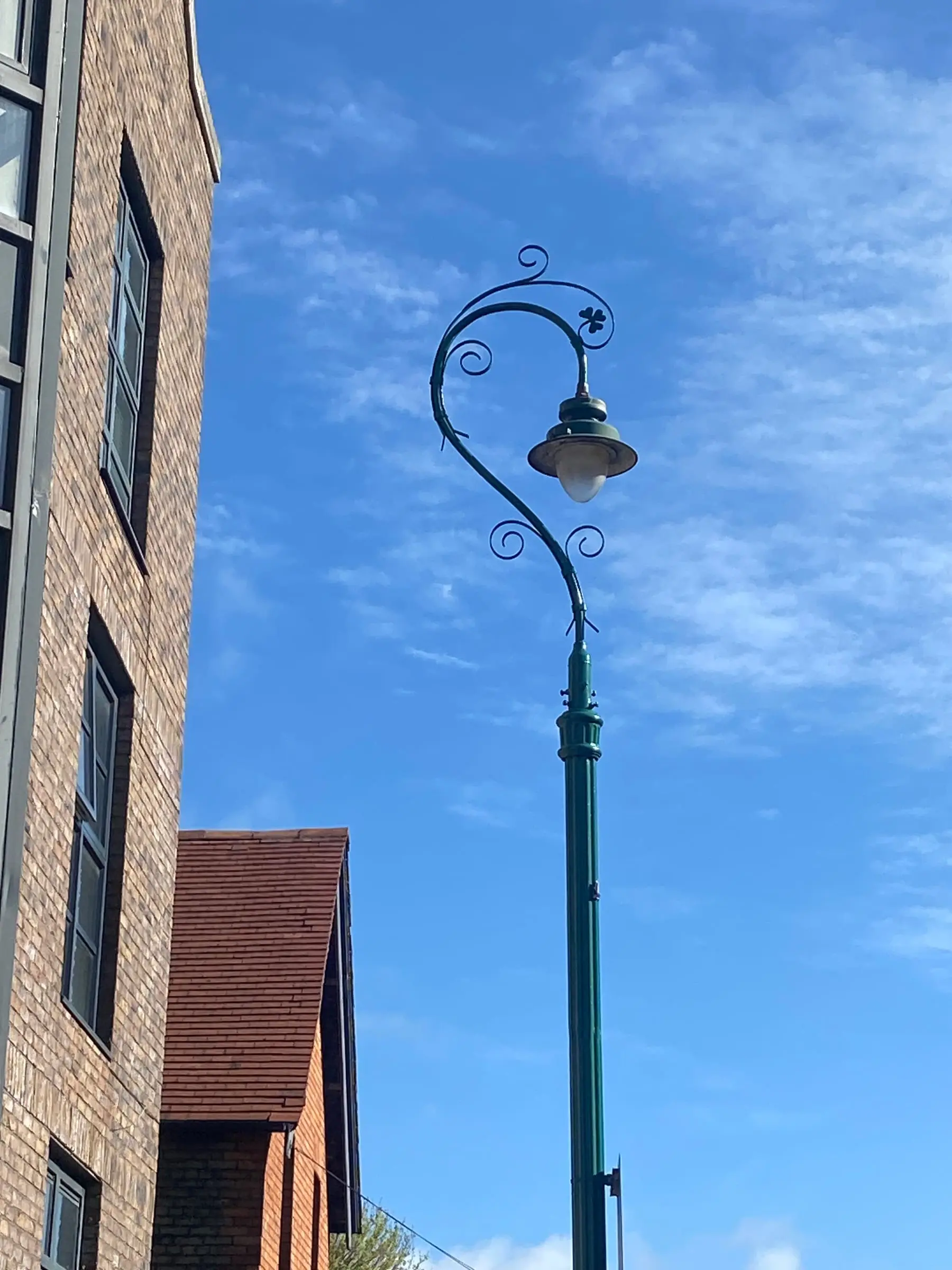 An ornate green cast-iron lamp post curling over a brick building under a blue sky with thin cloud