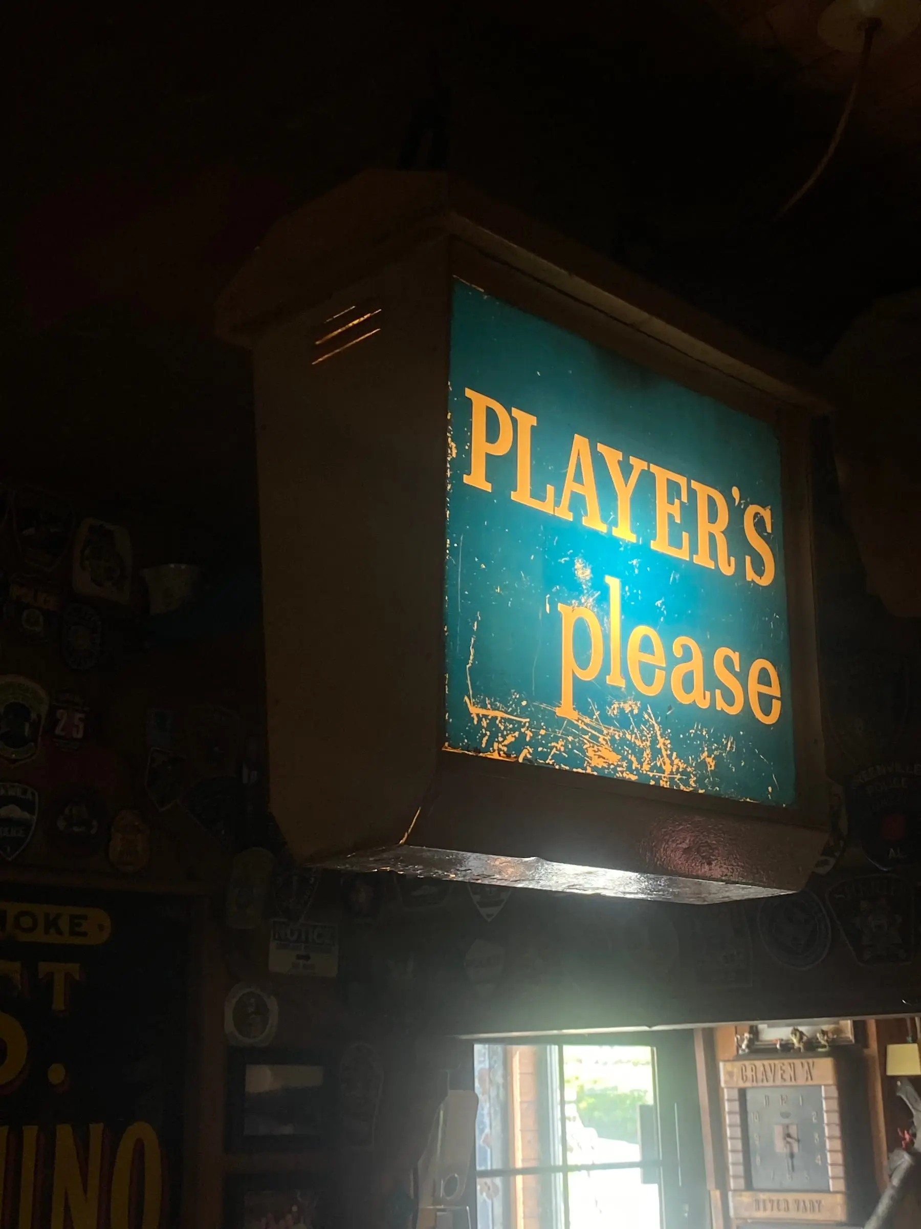 A hanging light box in a pub with orange letters on a teal background reading Player's please