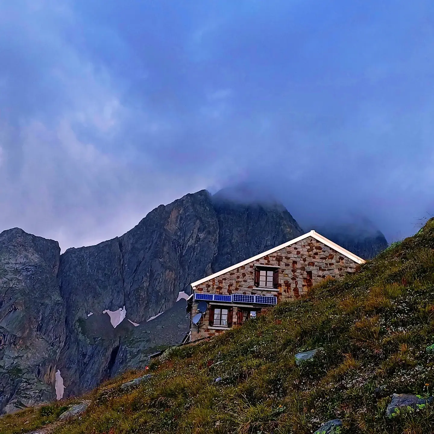 A stone mountain hut with solar panels on a grassy slope beneath a dark, cloud-wrapped peak